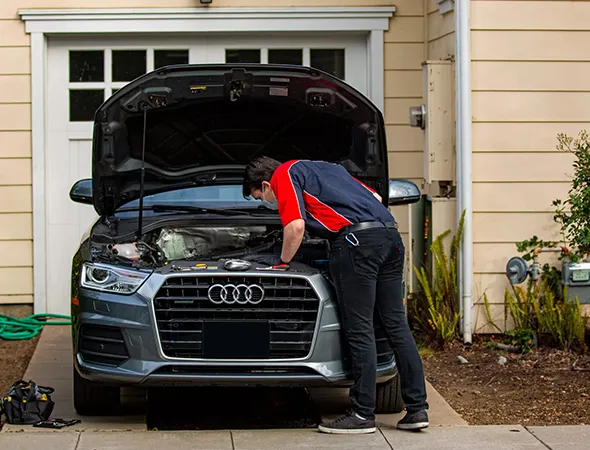 A mechanic in a red and navy uniform inspects the engine of a gray Audi parked in a driveway. The car's hood is open, and the scene is calm and focused.