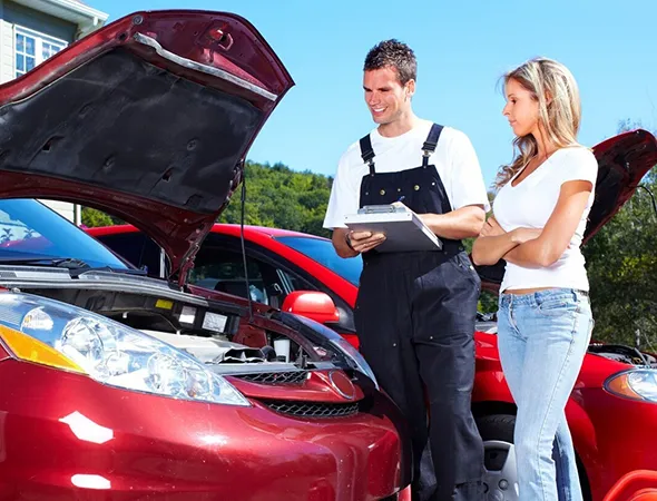 A mechanic with a clipboard inspects a red car's engine while a woman in casual attire watches, both appearing focused. Bright, sunny outdoor setting.