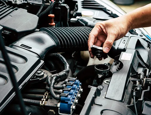 A hand unscrews the car's oil cap under the hood, surrounded by engine components and hoses. The scene conveys maintenance and care.