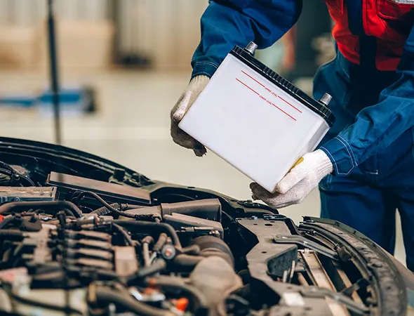 Mechanic in blue coveralls holding a white car battery, standing over an open car hood. The setting is a garage, conveying focus and professionalism.