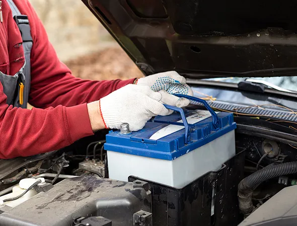 A person in a red sweater and gloves is installing a blue car battery under an open hood. The scene conveys a sense of maintenance and focus.