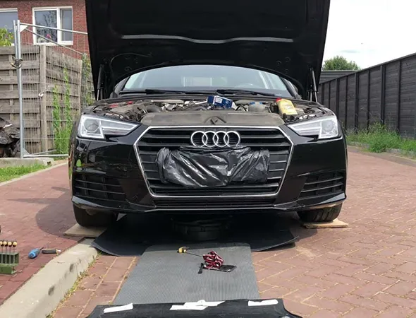 Black Audi car with open hood, parked on a driveway. Tools and a mat are on the ground, indicating it's being repaired or serviced. Calm, sunny day.