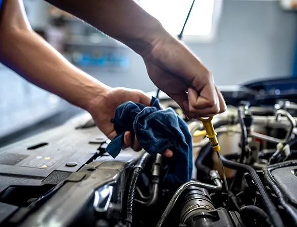 Hands check car oil under the open hood, using a dipstick and blue cloth. The scene conveys focus and maintenance in a garage setting.