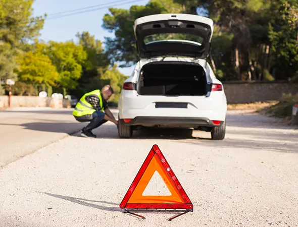 A person in a high-visibility vest inspects a flat tire on a parked car with an open trunk on a sunny road. A red warning triangle is placed nearby.