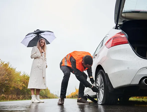A man in an orange vest changes a tire on a white car by a deserted road. A woman stands nearby with an umbrella, looking concerned. It's overcast and rainy.