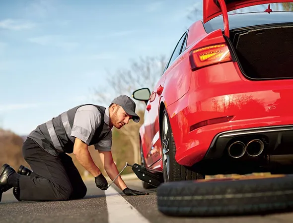 A man kneels beside a red car on the roadside, changing a tire. He wears a cap, gloves, and a safety vest. The image conveys urgency and focus.